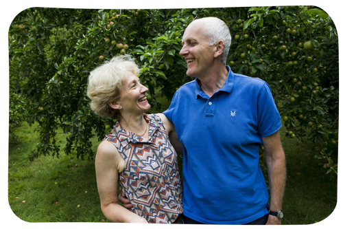 Happy older couple standing in a garden, with their arms around each other laughing together.